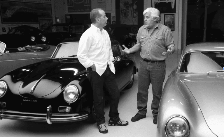 Two men standing and conversing in a garage surrounded by vintage cars, including a black and a silver Porsche. One man wears a white shirt and jeans, while the other sports a denim shirt and jeans.